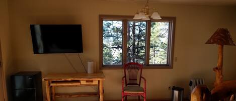 A room with a wooden desk, a red chair, a television mounted on the wall, and a window with a view of trees.