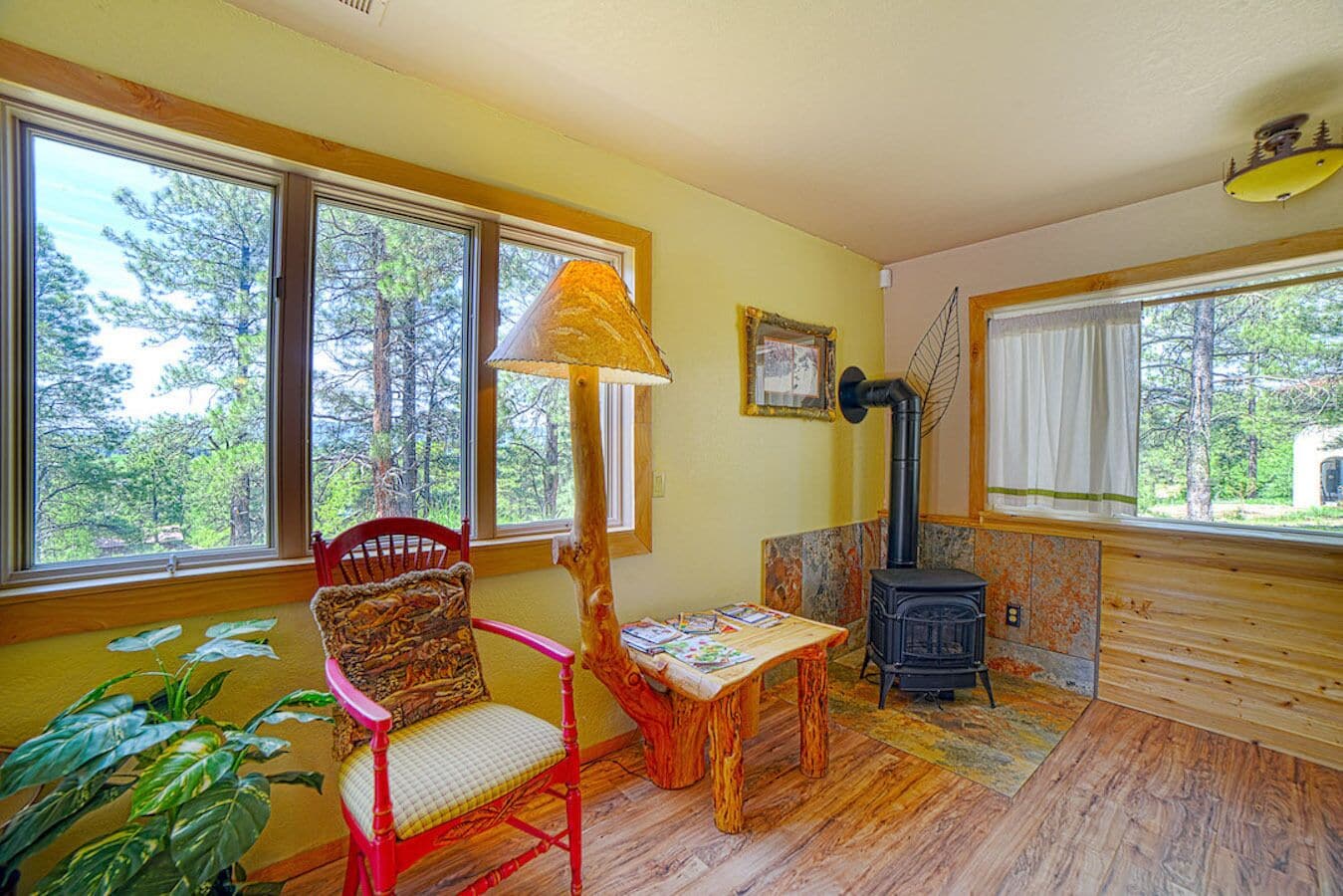 A cozy room with a wooden floor, a red chair, a table with books, a black wood stove, and large windows with a forest view.