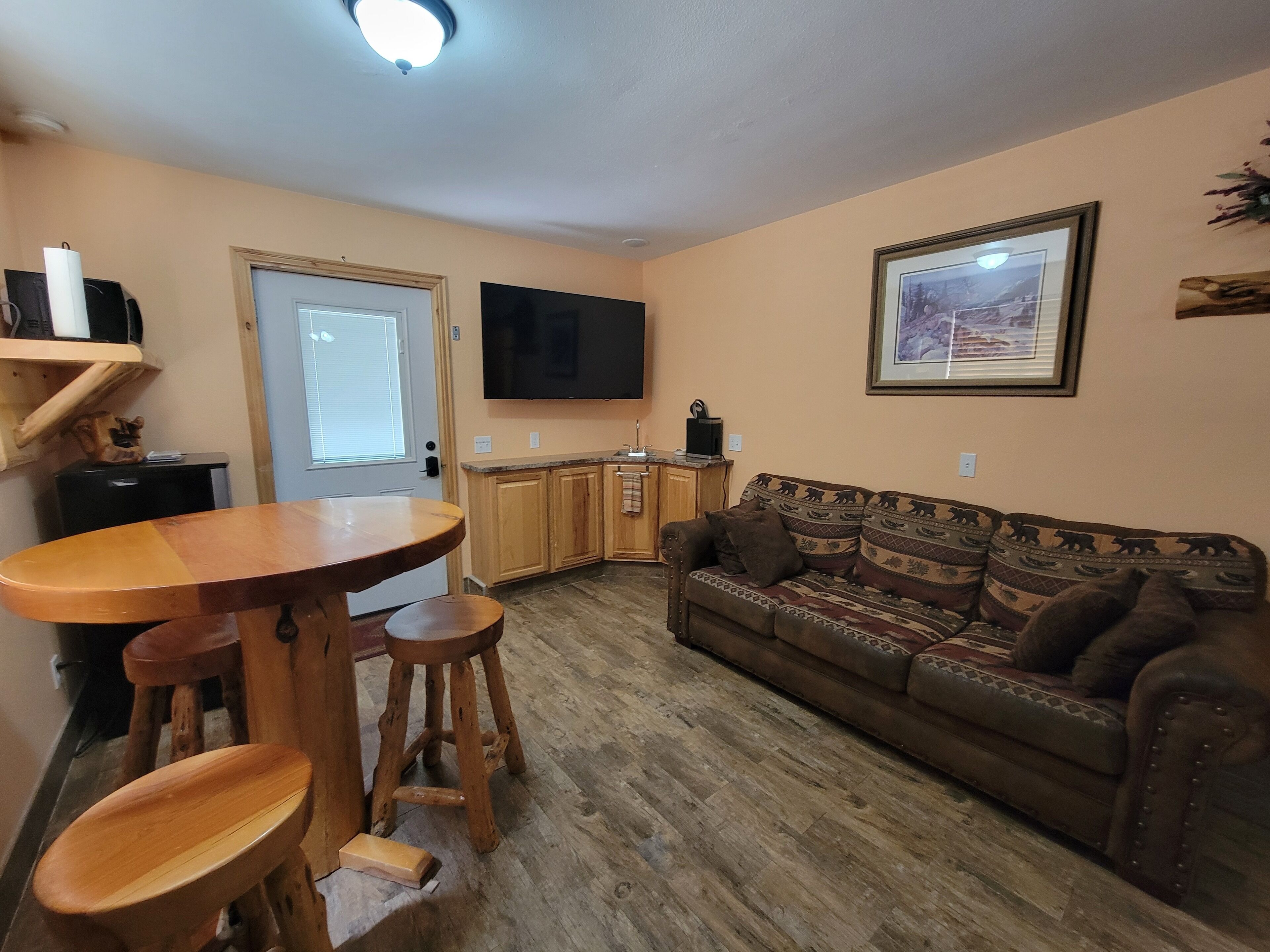 Una sala de estar con una mesa de comedor de madera, dos taburetes, un sofá y un televisor montado en la pared.