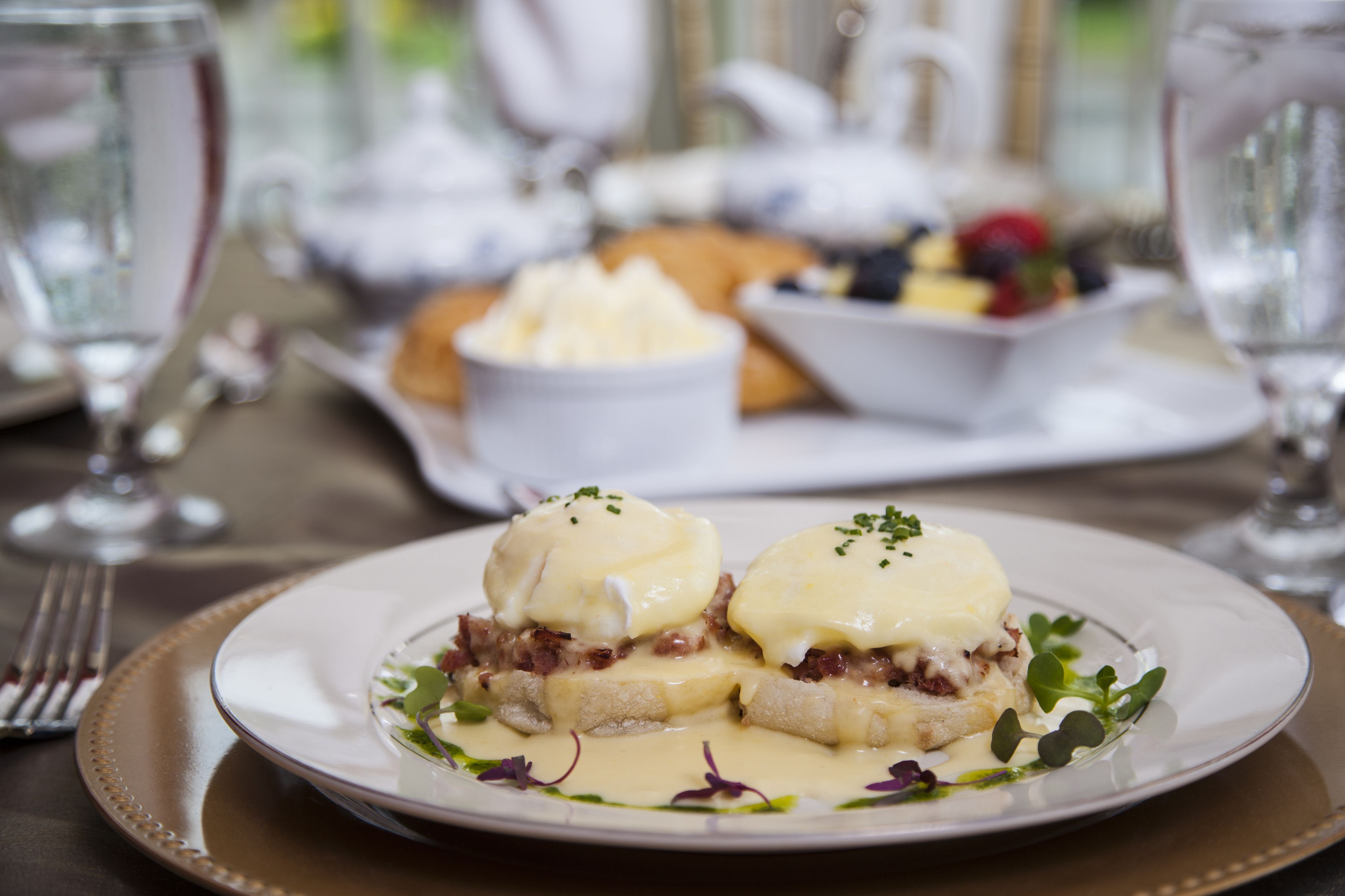 Une assiette d’œufs Bénédicte nappés de sauce hollandaise, accompagnée d’une portion de fruits et d’un petit ramequin de beurre.