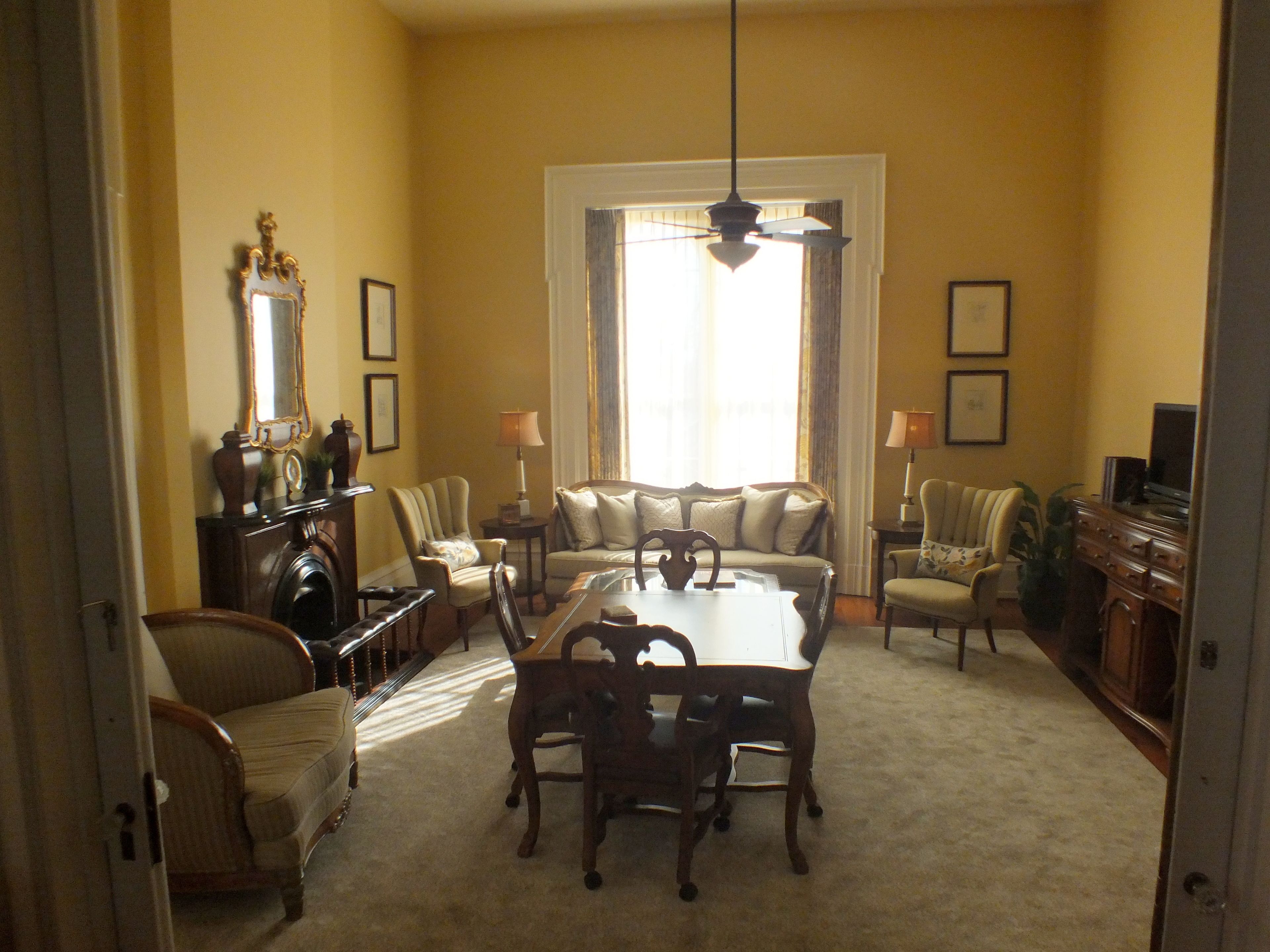 A dining room with a large table, chairs, and a chandelier. There are sofas and a television in the background.