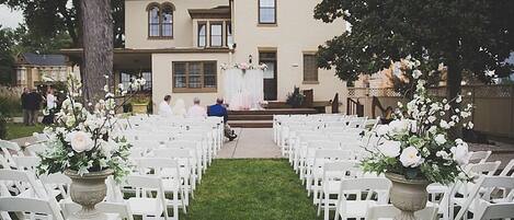 An outdoor wedding ceremony with white chairs arranged in rows, a stage with a floral backdrop, and a large house in the background.
