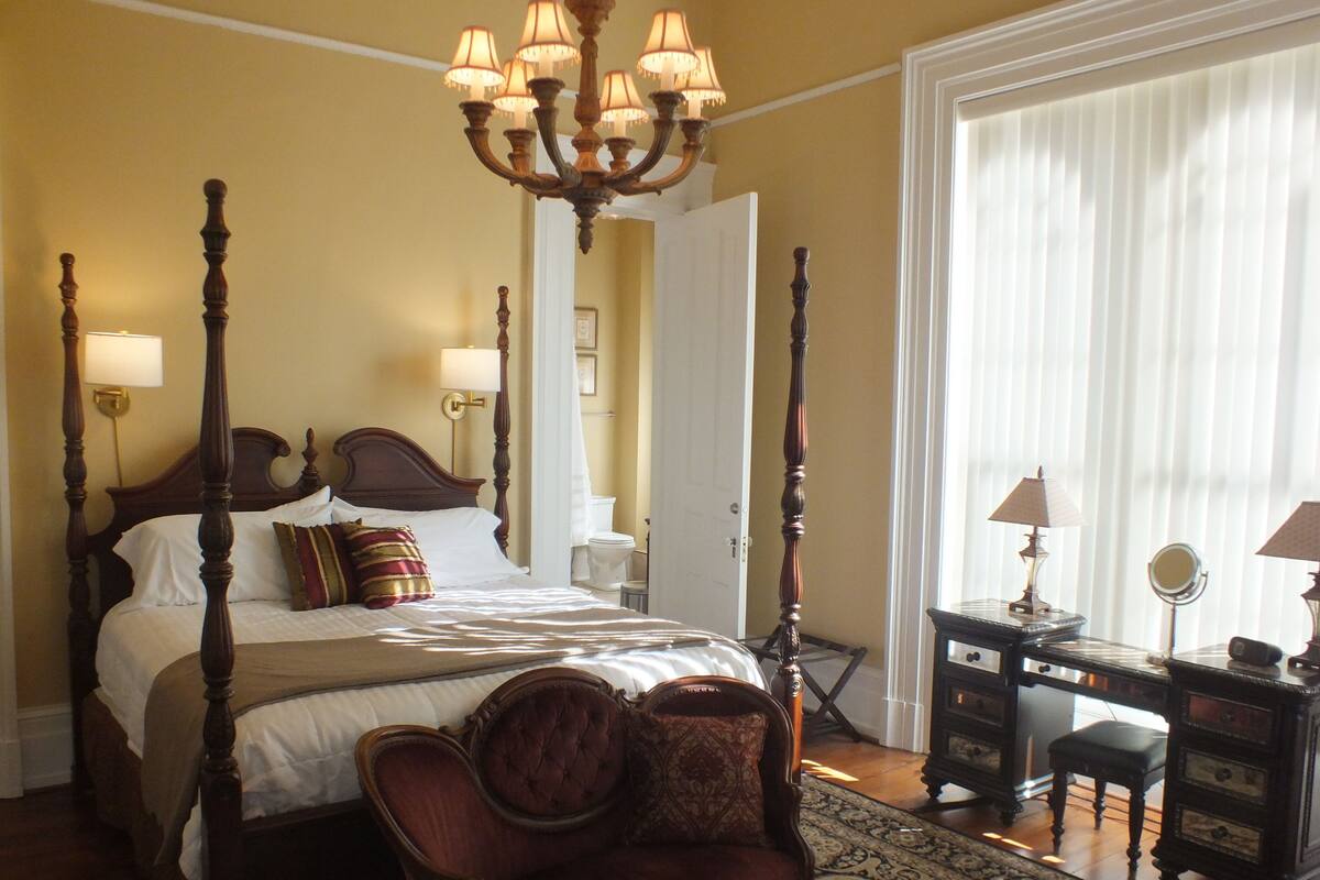 A four-poster bed with a chandelier, a wooden desk with a mirror, and a patterned rug.