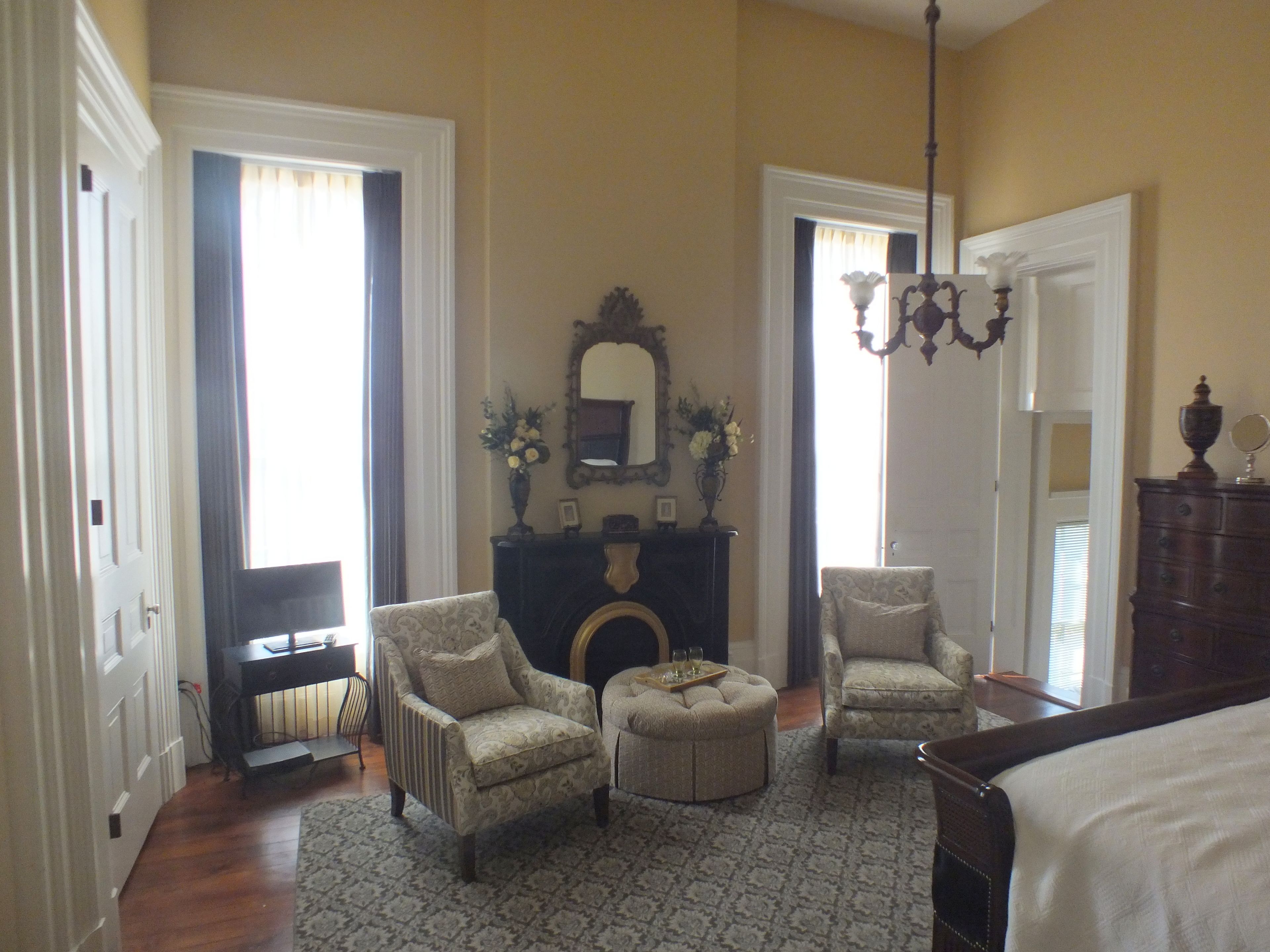 A spacious bedroom with a large bed, two armchairs, a small ottoman, a fireplace, and a chandelier.