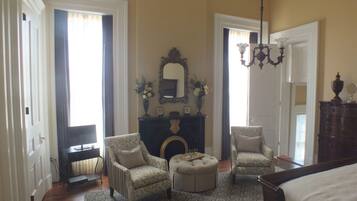A spacious bedroom with a large bed, two armchairs, a small ottoman, a fireplace, and a chandelier.