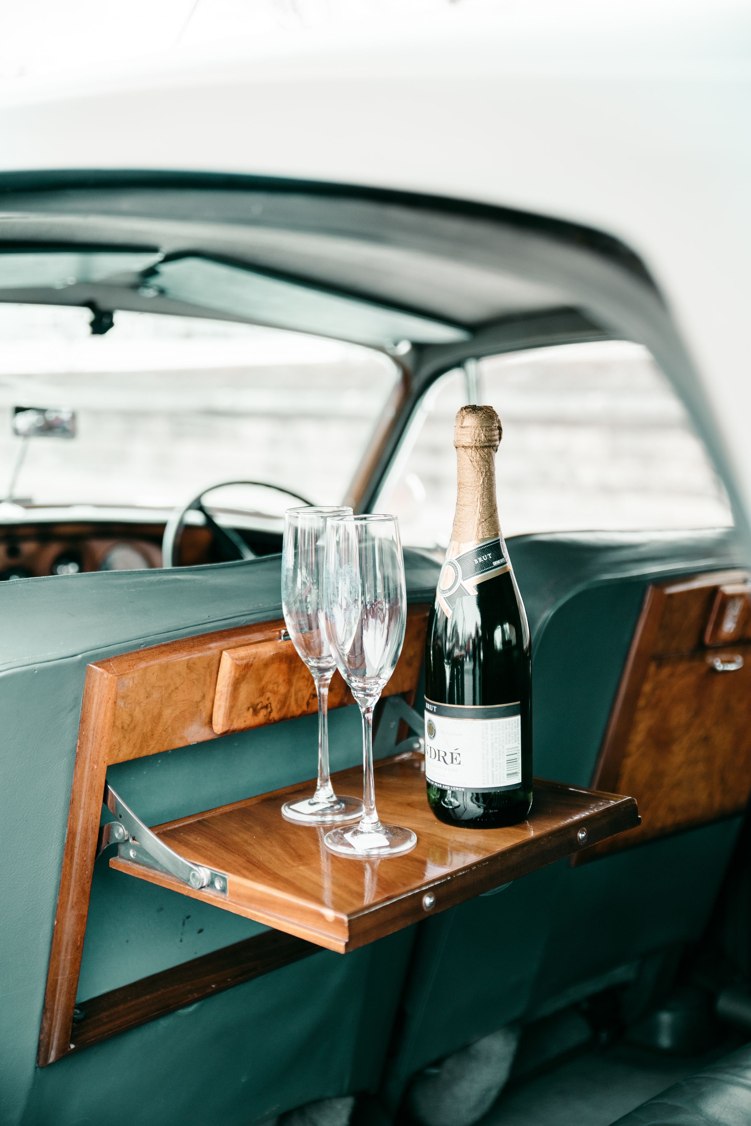 A bottle of champagne and two glasses on a wooden tray in a car's interior.