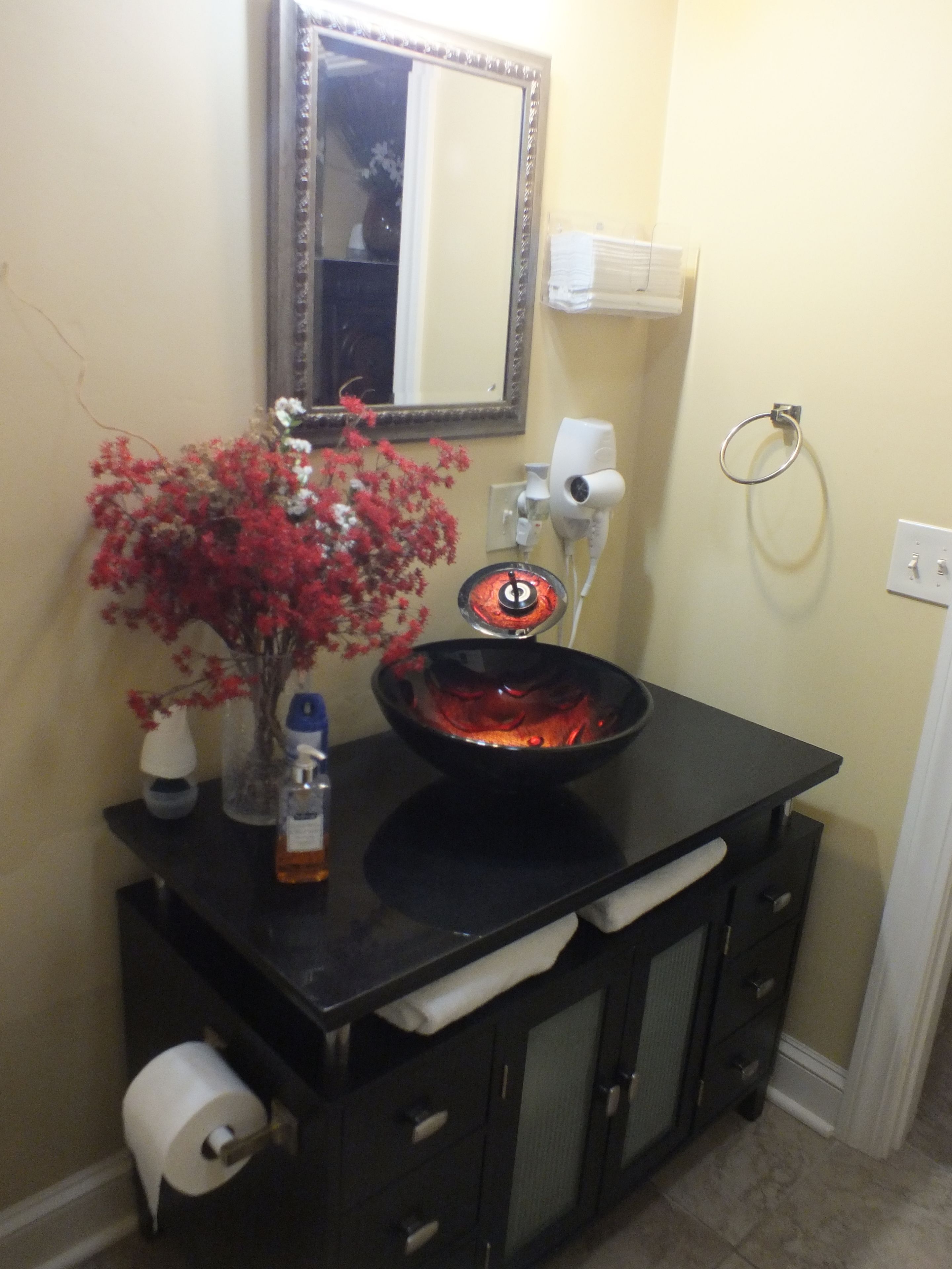A bathroom with a black vanity, a glass bowl sink, a mirror, a hairdryer, a vase with red flowers, and a toilet paper holder.