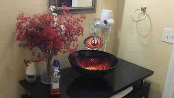 A bathroom with a black vanity, a glass bowl sink, a mirror, a hairdryer, a vase with red flowers, and a toilet paper holder.