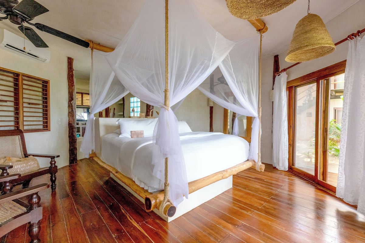 Un dormitorio amplio con una cama grande, suelo de madera, ventilador de techo y vistas al exterior a través de un ventanal.