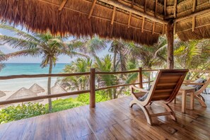 A wooden deck with a thatched roof overlooking a beach with palm trees and a wooden railing.