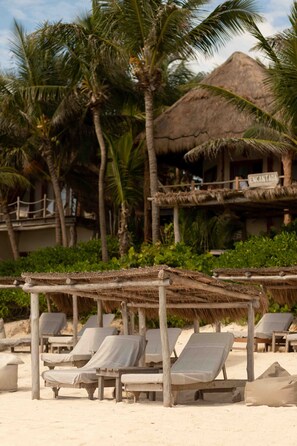 A beach with thatched-roof structures, palm trees, and lounge chairs.
