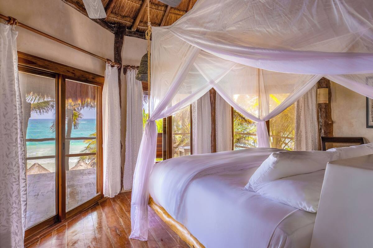 Un dormitorio con una cama grande, suelo de madera y vista a la playa a través de una ventana.