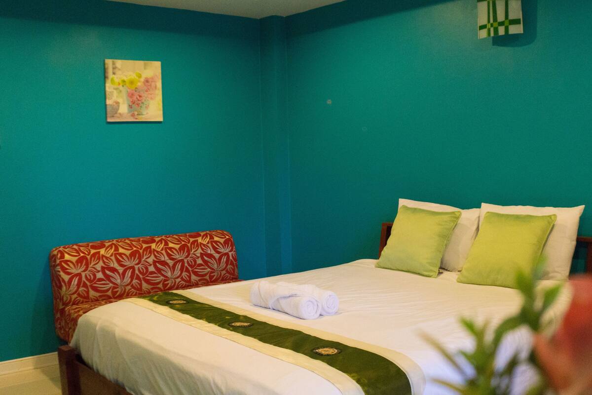 Habitación de hotel con paredes color turquesa, cama con almohadas verdes y blancas, cabecera estampada con motivos florales y un cuadro en la pared.