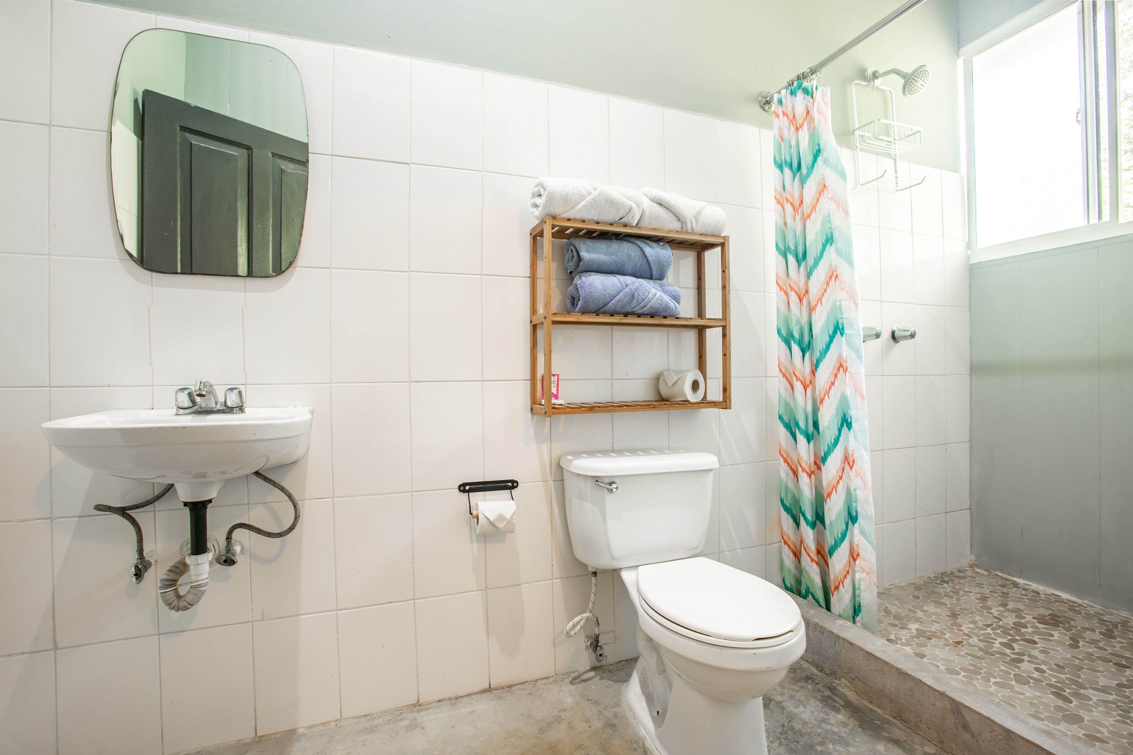 A bathroom with a white sink, toilet, and shower. There is a mirror above the sink and a shelf with towels.
