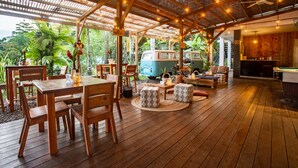 Un espacio exterior cubierto con suelo de madera, una mesa de comedor con sillas, una furgoneta azul y una mesa de billar.
