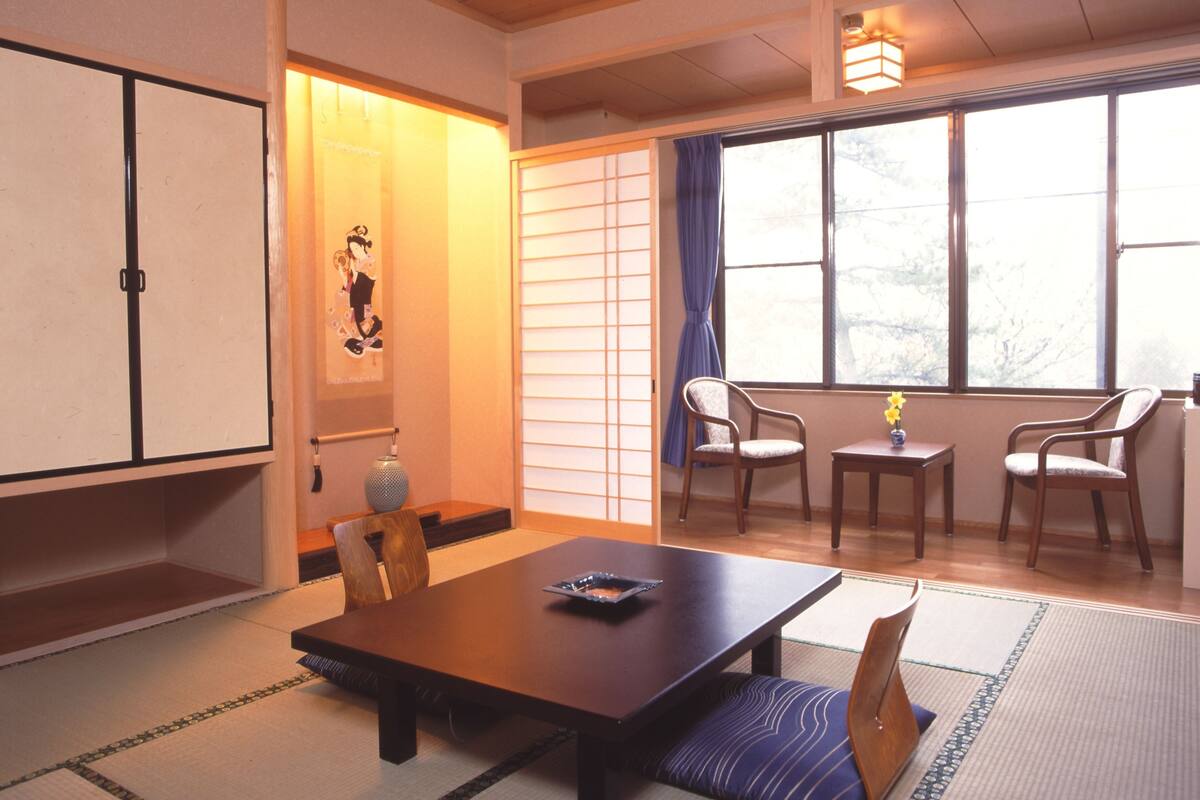 A traditional Japanese room with tatami flooring, a low wooden table, two chairs, a small table with a vase, and a sliding door with a decorative panel.