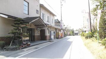 Vista de una calle con un edificio de estilo arquitectónico tradicional japonés, con tejado de tejas y elementos de madera. Hay carteles en japonés, y la calle está bordeada de árboles y arbustos.