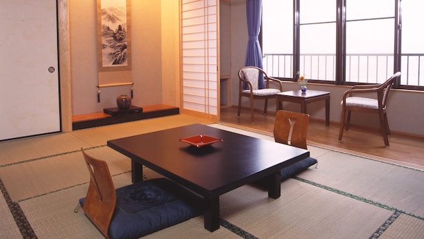 Une pièce de style japonais traditionnel, avec un sol recouvert de tatamis, une petite table basse avec des coussins, une chaise en bois et une porte coulissante donnant sur l’extérieur.