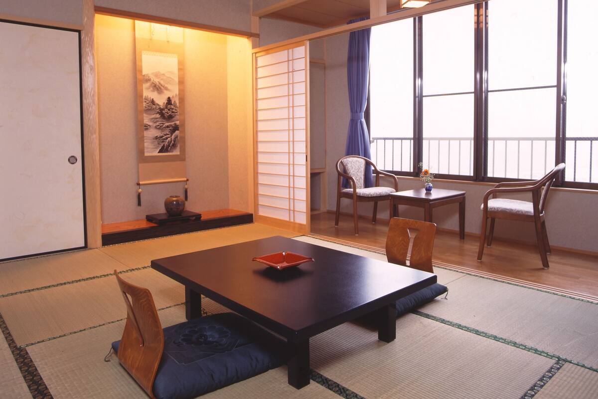A traditional Japanese room with tatami flooring, a low table with cushions, a wooden chair, and a sliding door with a view of the outside.