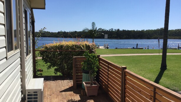 A wooden deck with a view of a lake, a grassy area, and a wooden fence. - Riviera Caravan Park (Sussex Inlet)