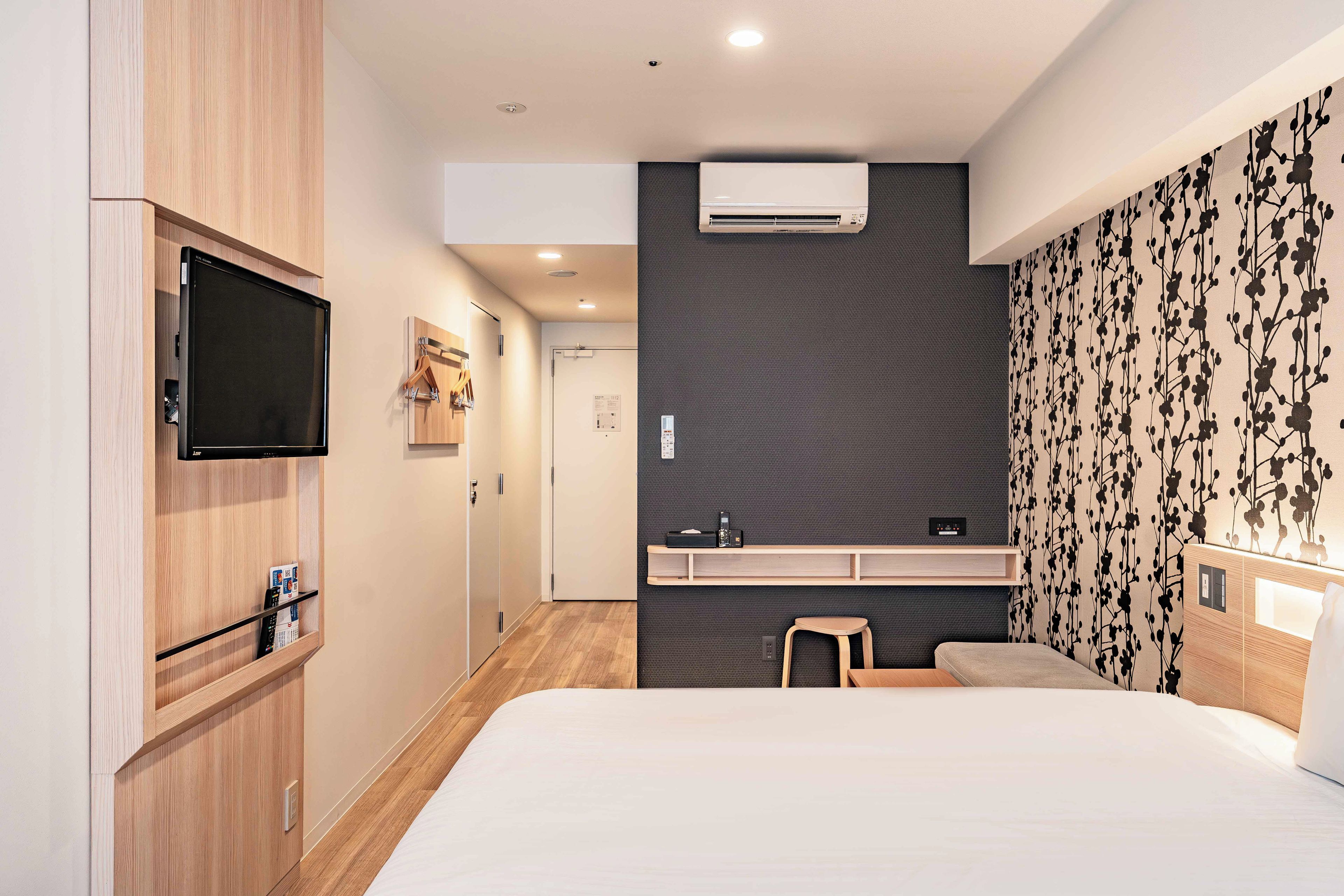 A modern hotel room with a bed, a TV mounted on the wall, a small desk with a chair, and an air conditioning unit.