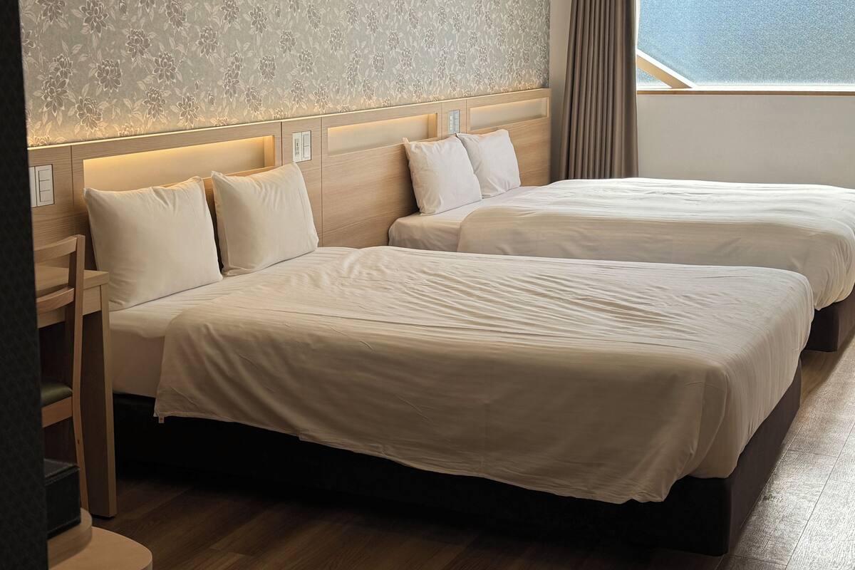 Ein Hotelzimmer mit zwei Betten, einem Holzkopfteil, einem Nachttisch und einem Fenster mit Vorhängen.