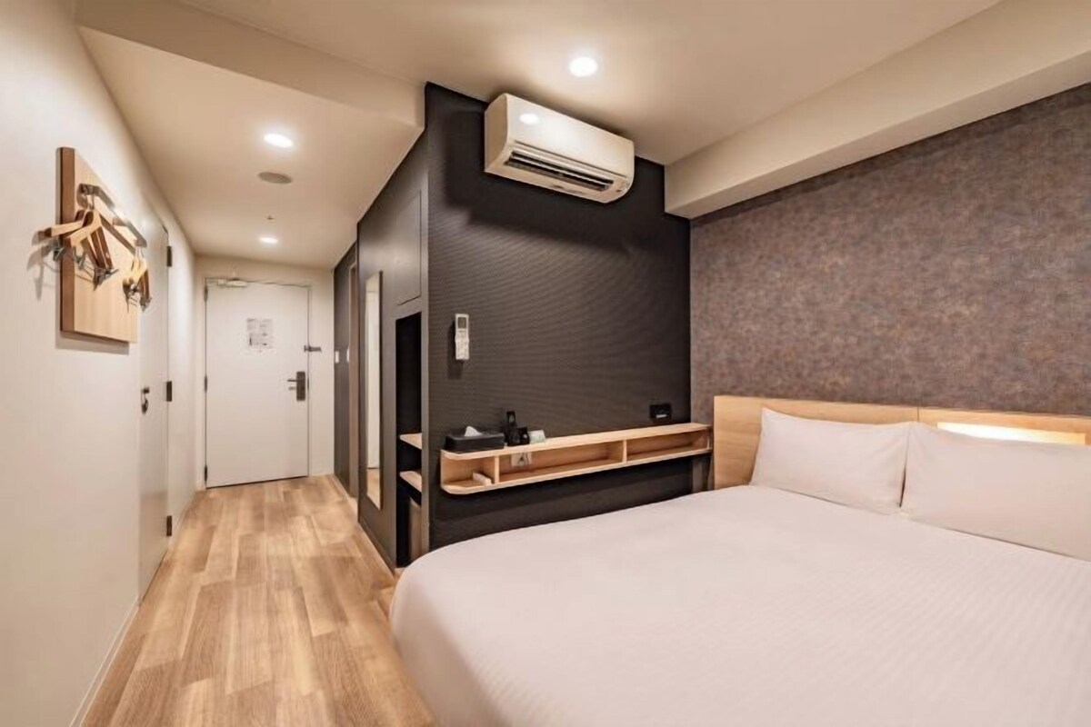 Una habitación de hotel con cama, mesita de noche, equipo de aire acondicionado y suelo de madera.