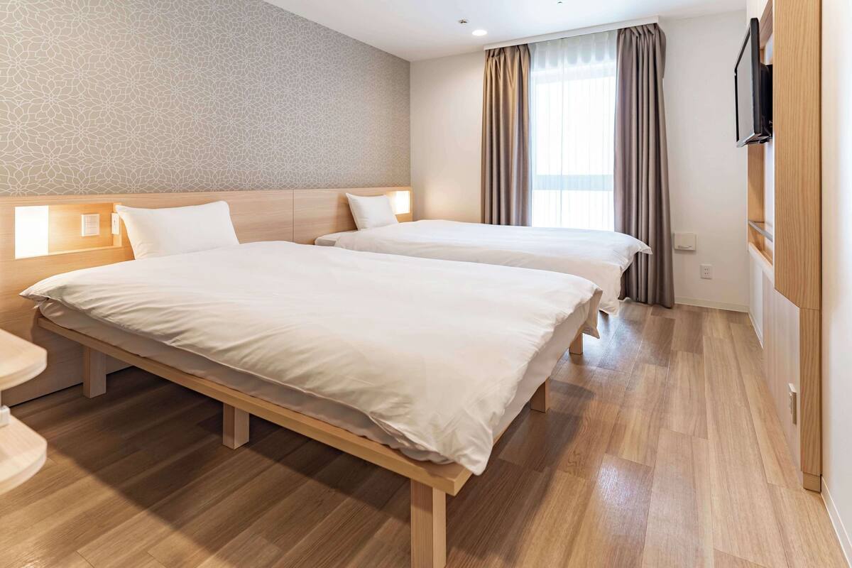 Ein Hotelzimmer mit zwei Betten, Holz-Kopfteilen, einem Flachbildfernseher und einem großen Fenster mit Vorhängen.