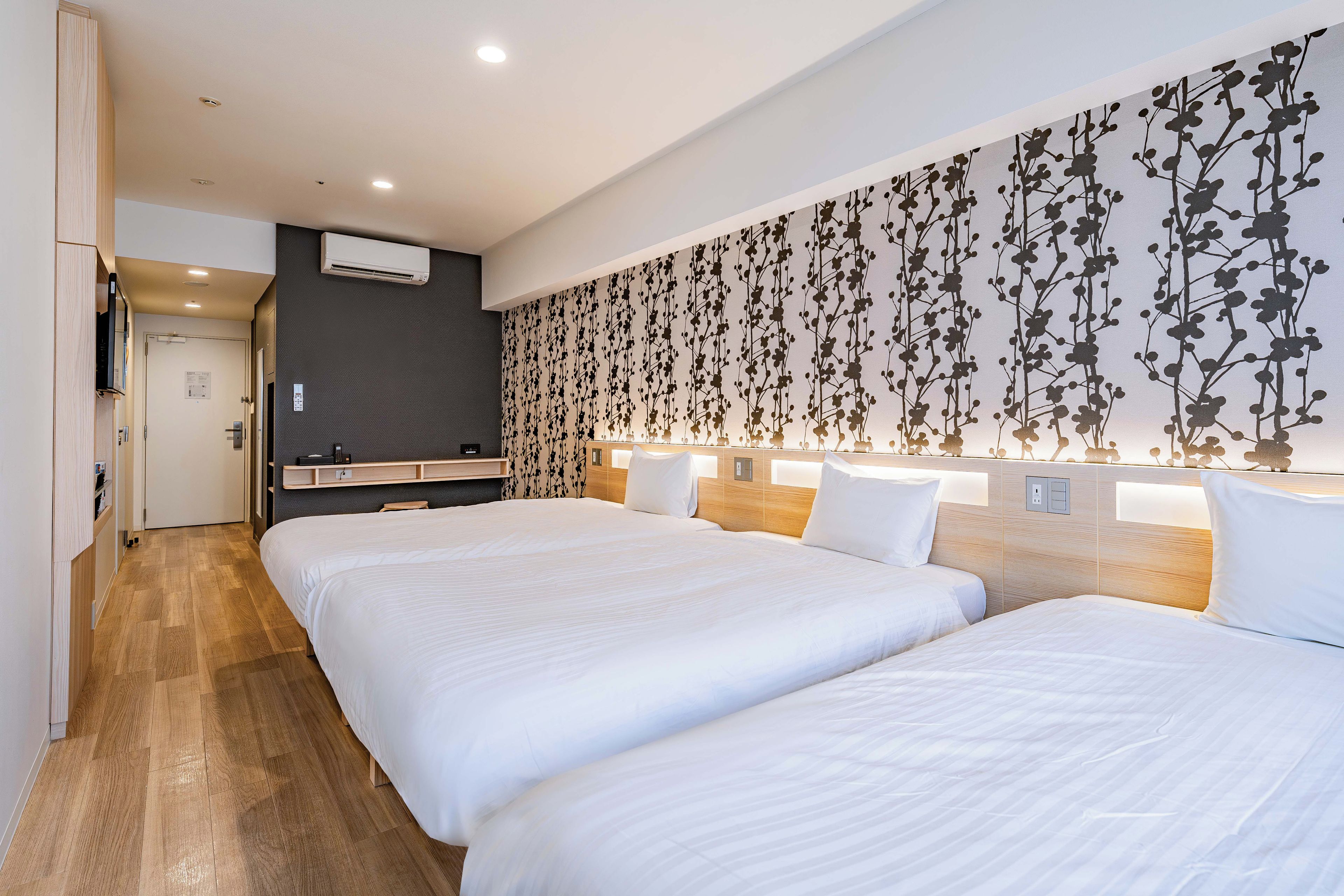 A hotel room with two beds, a wooden floor, a dark wall with a floral pattern, and a small kitchenette area.