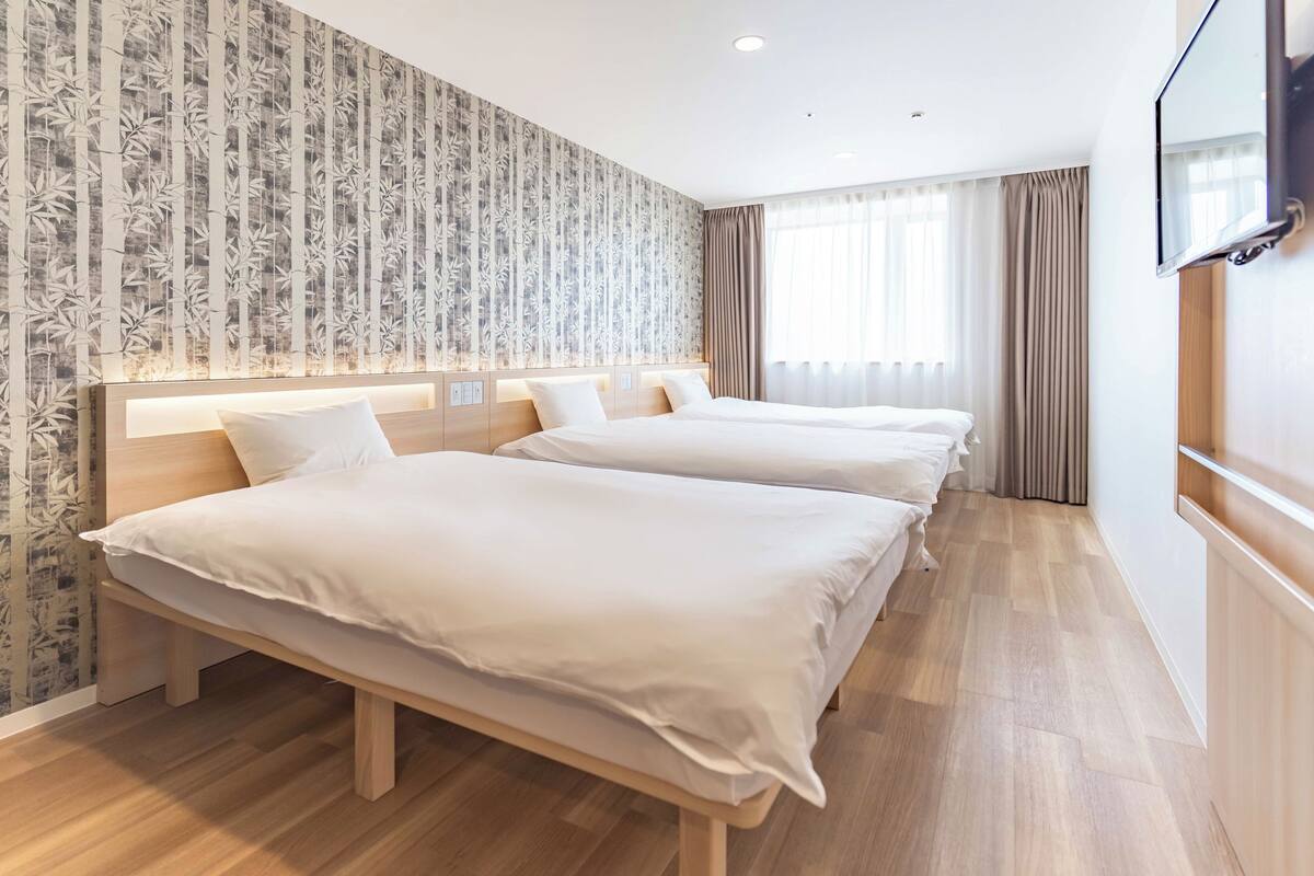 Ein Hotelzimmer mit zwei Betten, einem großen Fenster mit Vorhängen, einem an der Wand befestigten Fernseher und einem Wandpapier mit Muster.