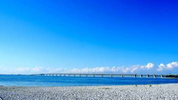 A pebbled beach with a clear blue sky and a long bridge in the distance.