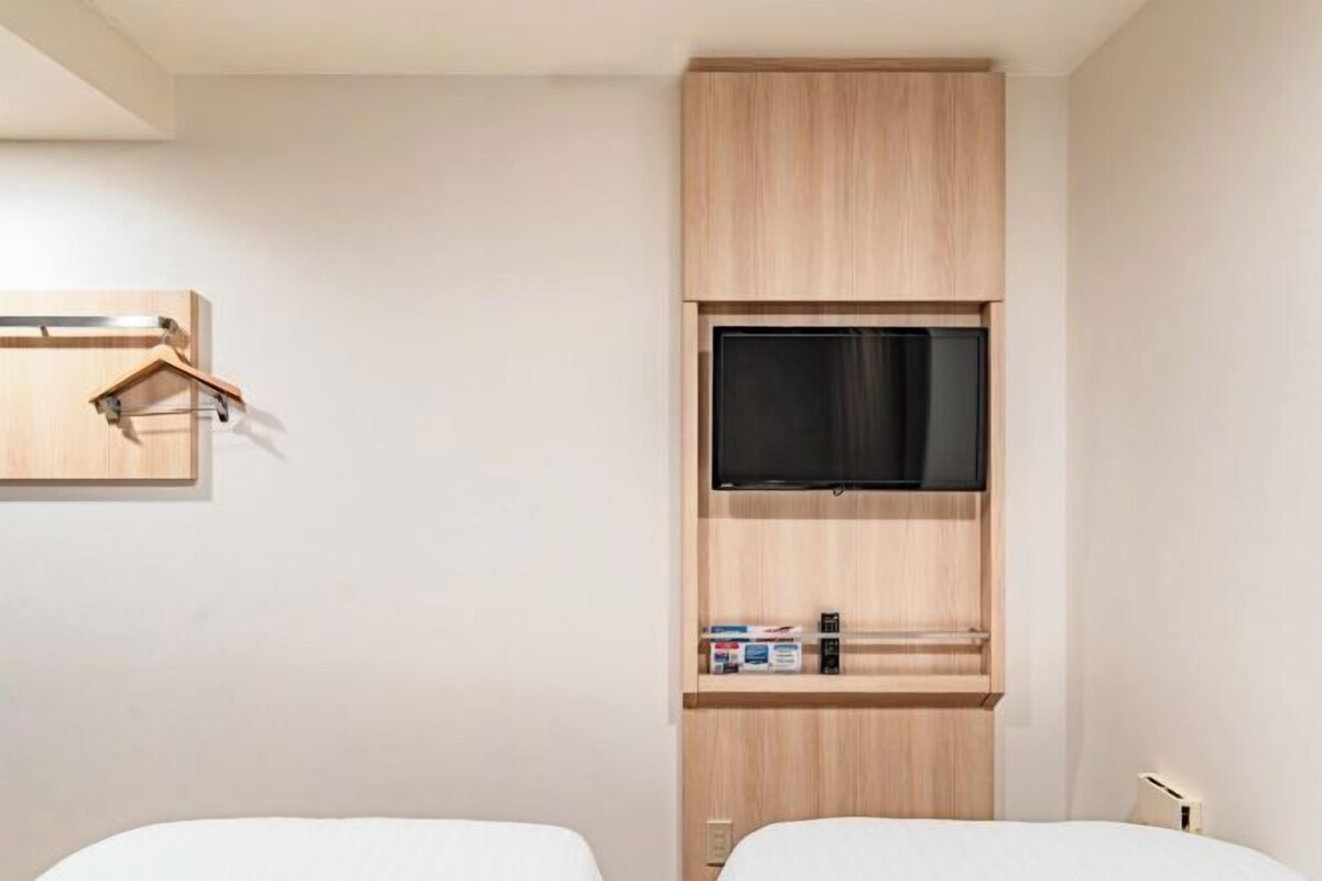 A hotel room with a bed, a TV mounted on a wooden cabinet, and a small shelf with items.