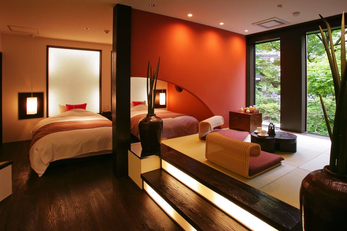 Ett modernt hotellrum med två sängar, en sittgrupp med en bänk, en vas och utsikt över grönområden utanför.
