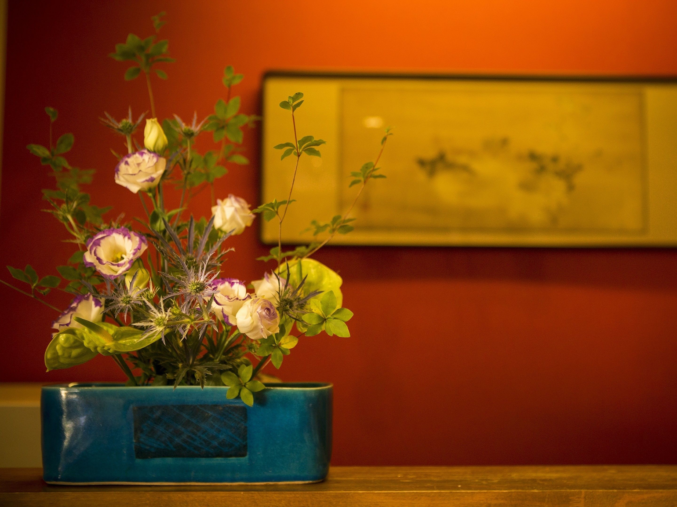 A blue vase with a floral arrangement on a wooden surface against a red wall with a framed picture.