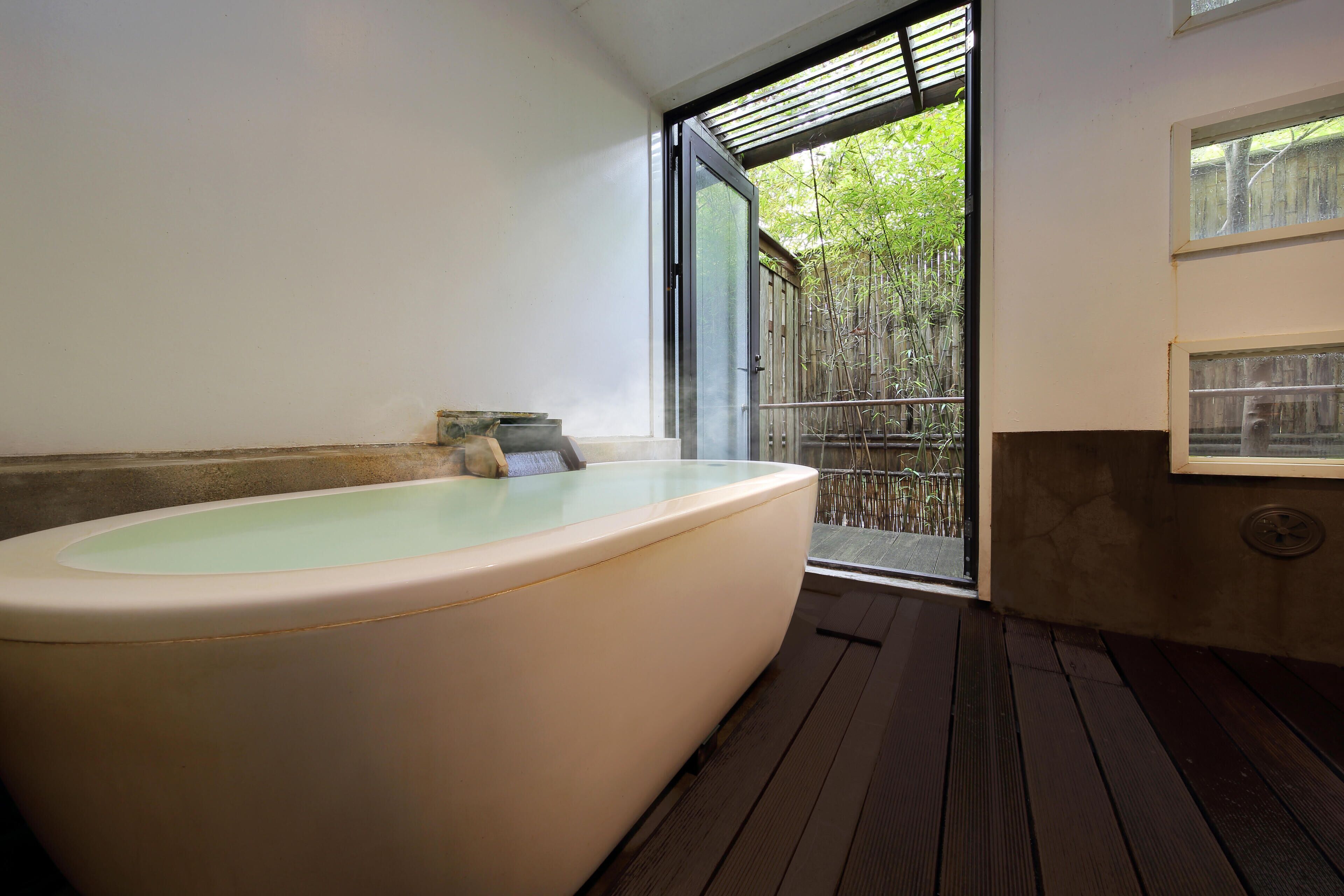 A modern bathroom with a large bathtub, a wooden floor, and a sliding glass door leading to an outdoor area.
