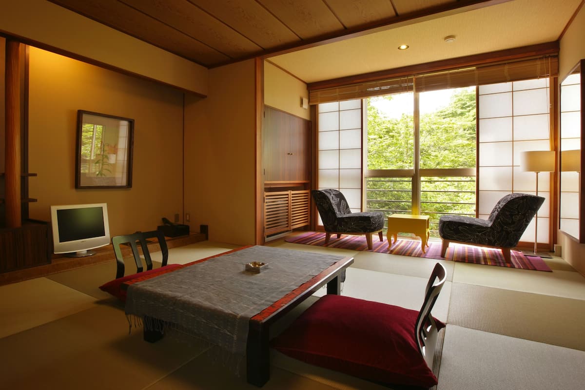 Ett traditionellt japanskt rum med tatamimattor på golvet, ett lågt bord med en röd kudde, en tv och en skjutdörr som leder ut till en balkong med stolar.