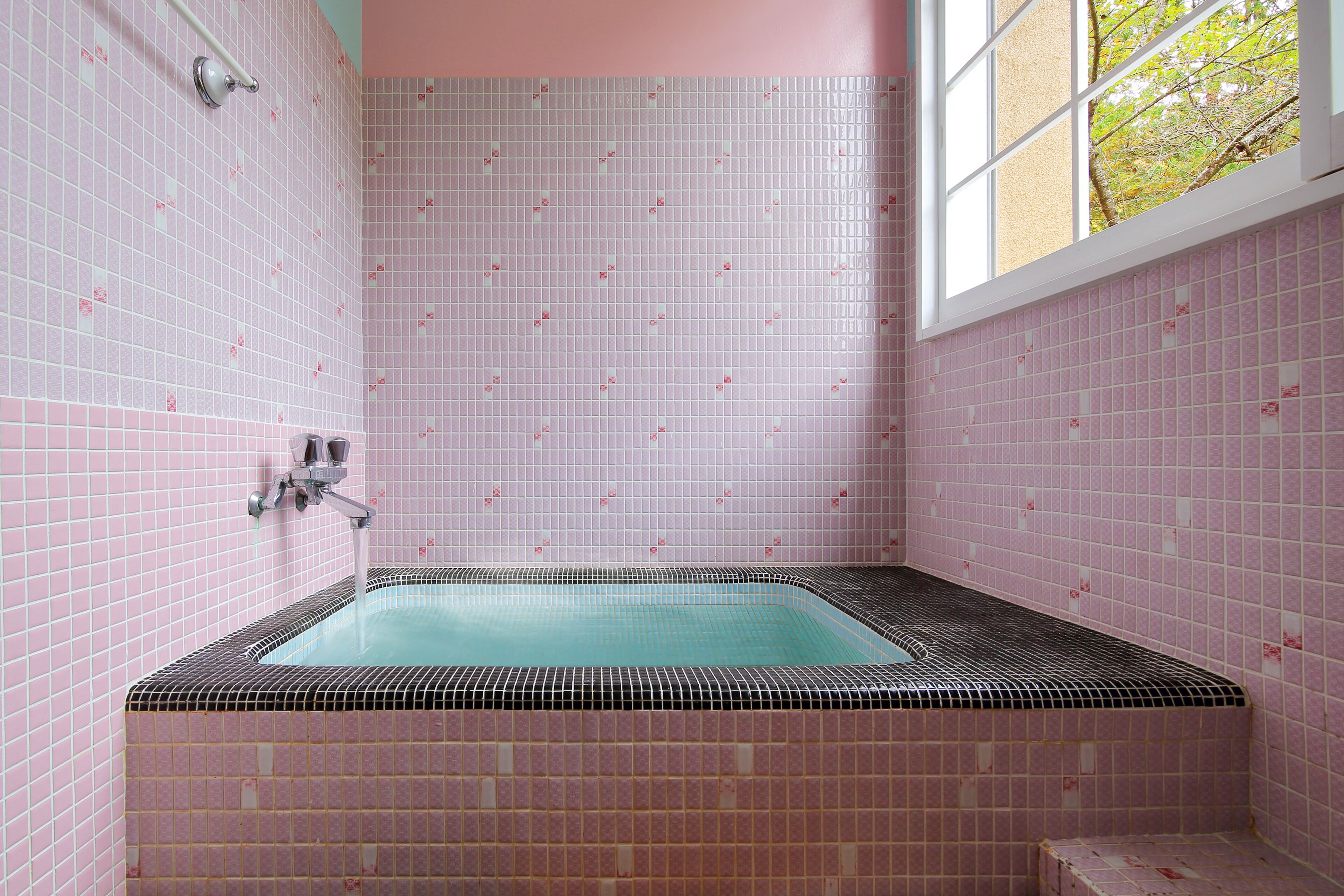 A bathroom with pink tiled walls, a bathtub, and a window with a view of trees.
