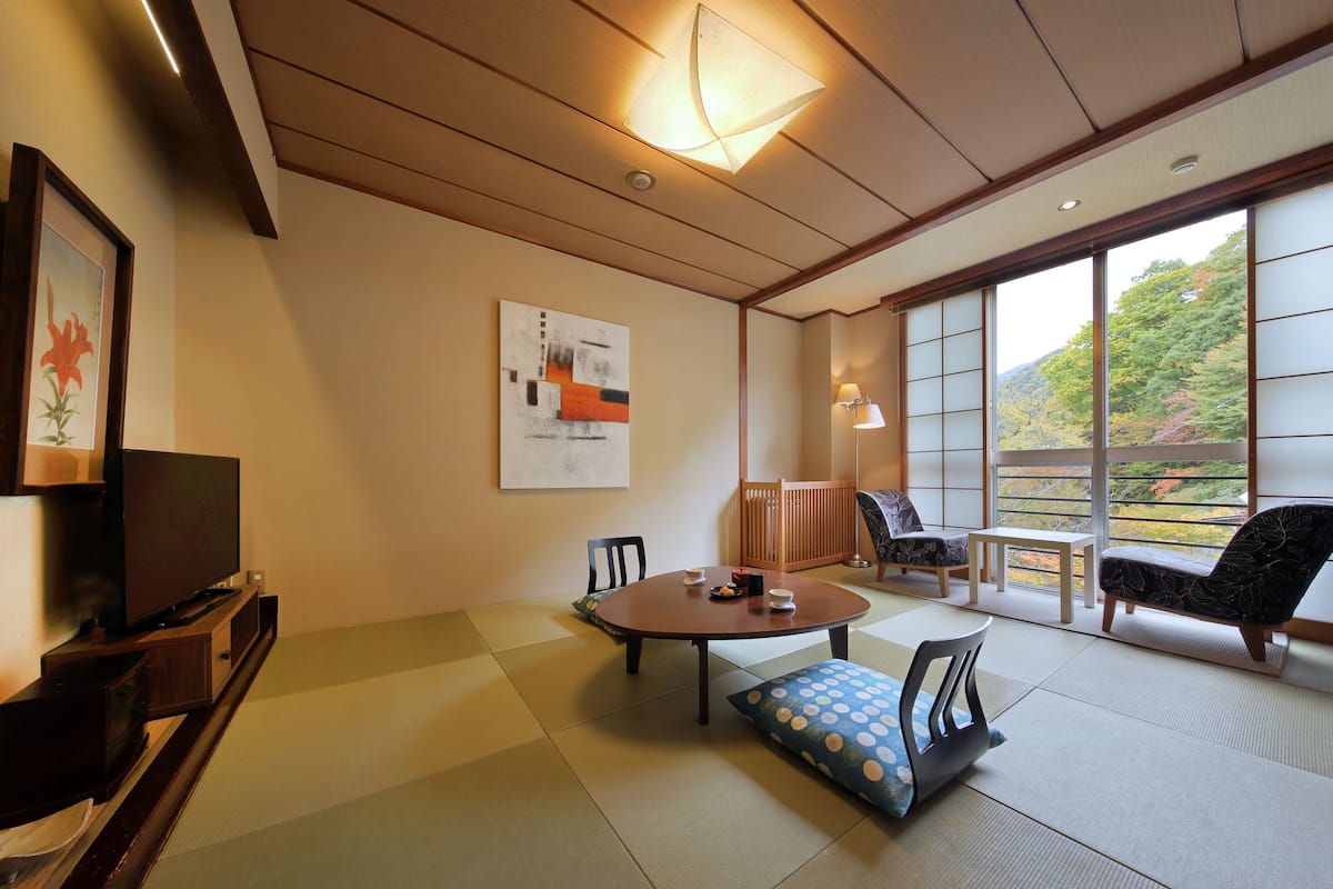 Ett traditionellt japanskt rum med tatamimattor på golvet, en tv, en tavla och utsikt över en skog genom stora fönster.