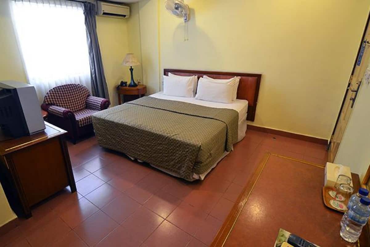 Une chambre d’hôtel avec un lit, une chaise, un bureau avec une télévision, une table de chevet avec une lampe et une bouteille d’eau posée sur le sol.