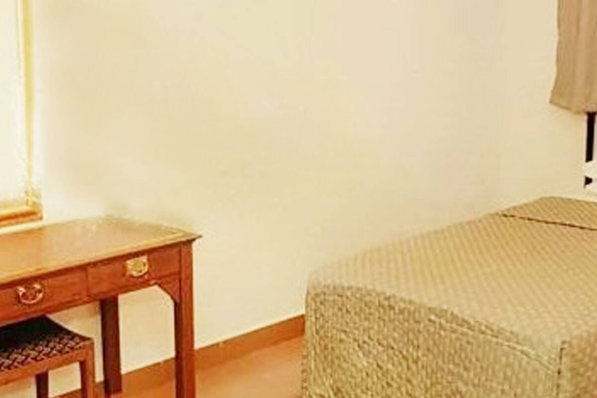 Un petit lit bien fait, avec un oreiller blanc et une couverture beige, une table de chevet en bois avec un tiroir, et une lampe sur une petite table placée à côté du lit.