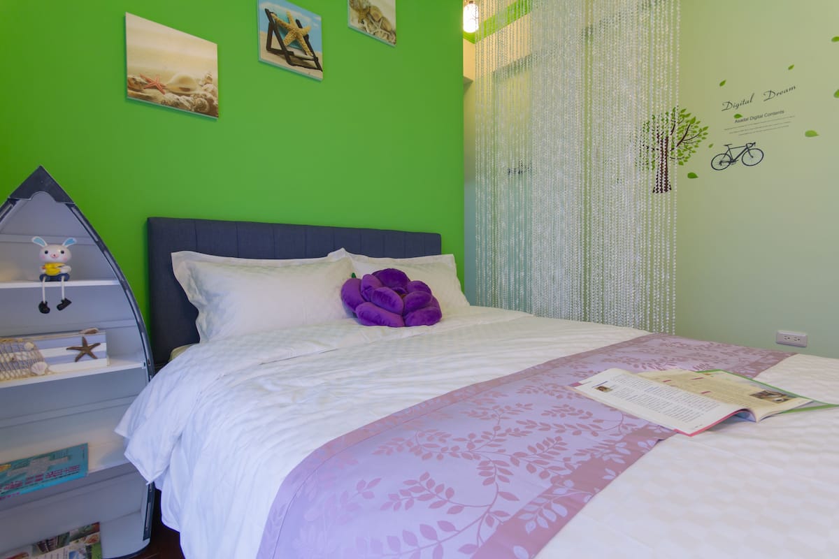 A bedroom with a green wall, a bed with a white and purple floral bedspread, a purple pillow, and a shelf with toys and books.