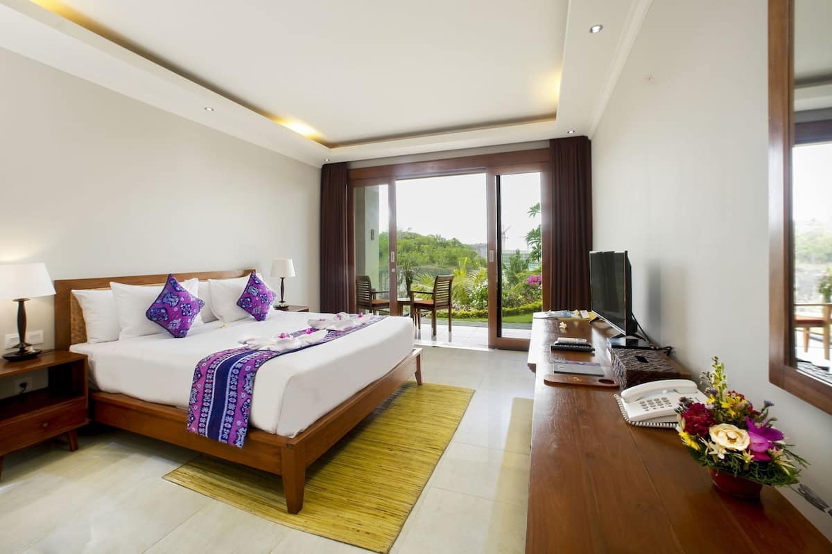 A hotel room with a bed, bedside tables, a desk with a TV, a phone, and a view of greenery.