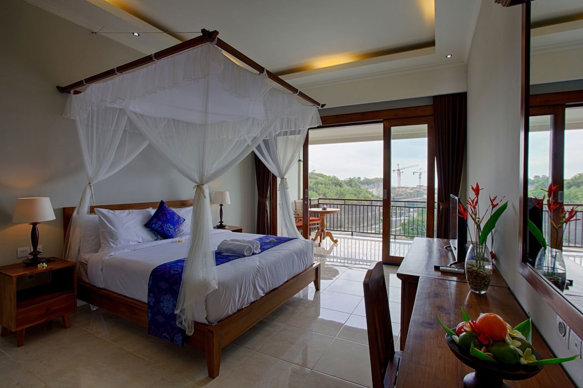 A four-poster bed with a canopy, a wooden nightstand with a lamp, a dining table with chairs, and a view of a balcony with a railing.