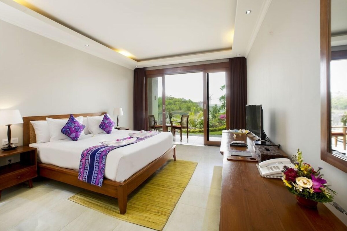 A hotel room with a bed, bedside table, lamp, TV, desk, chair, and a view of greenery.