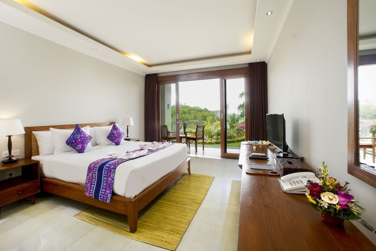 A hotel room with a bed, bedside tables, a desk with a TV, a phone, and a view of greenery outside.