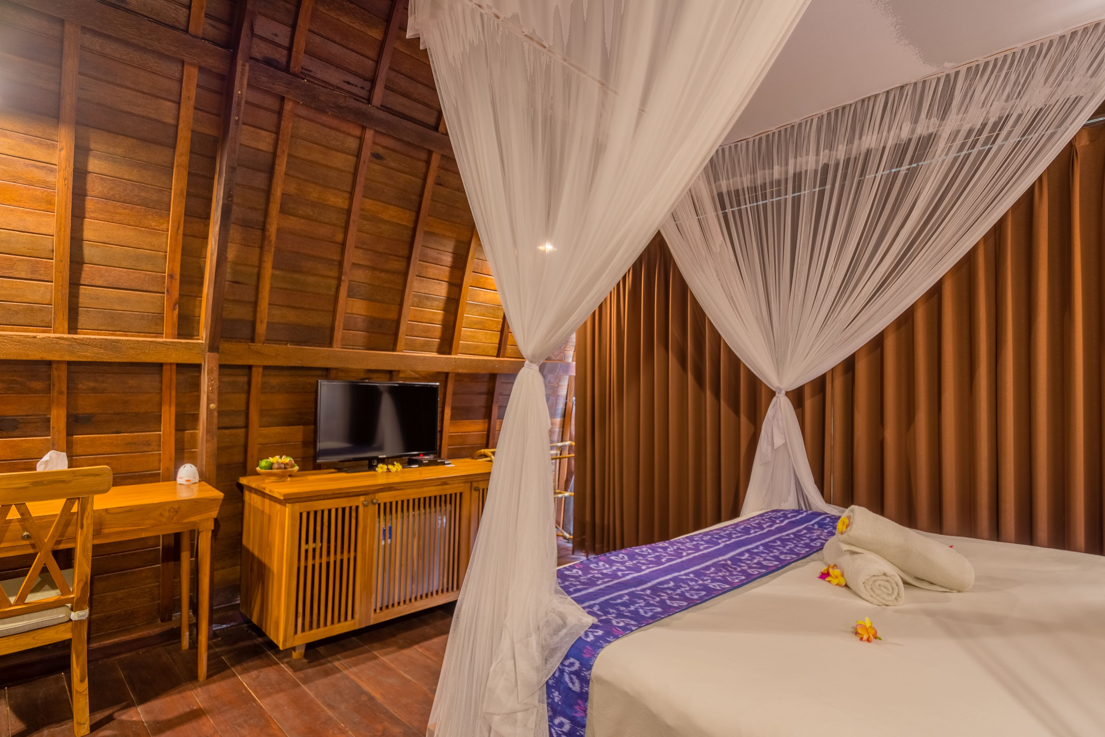 Une pièce avec un plafond en bois, un lit à baldaquin, un téléviseur posé sur un meuble en bois et un bureau avec une chaise.