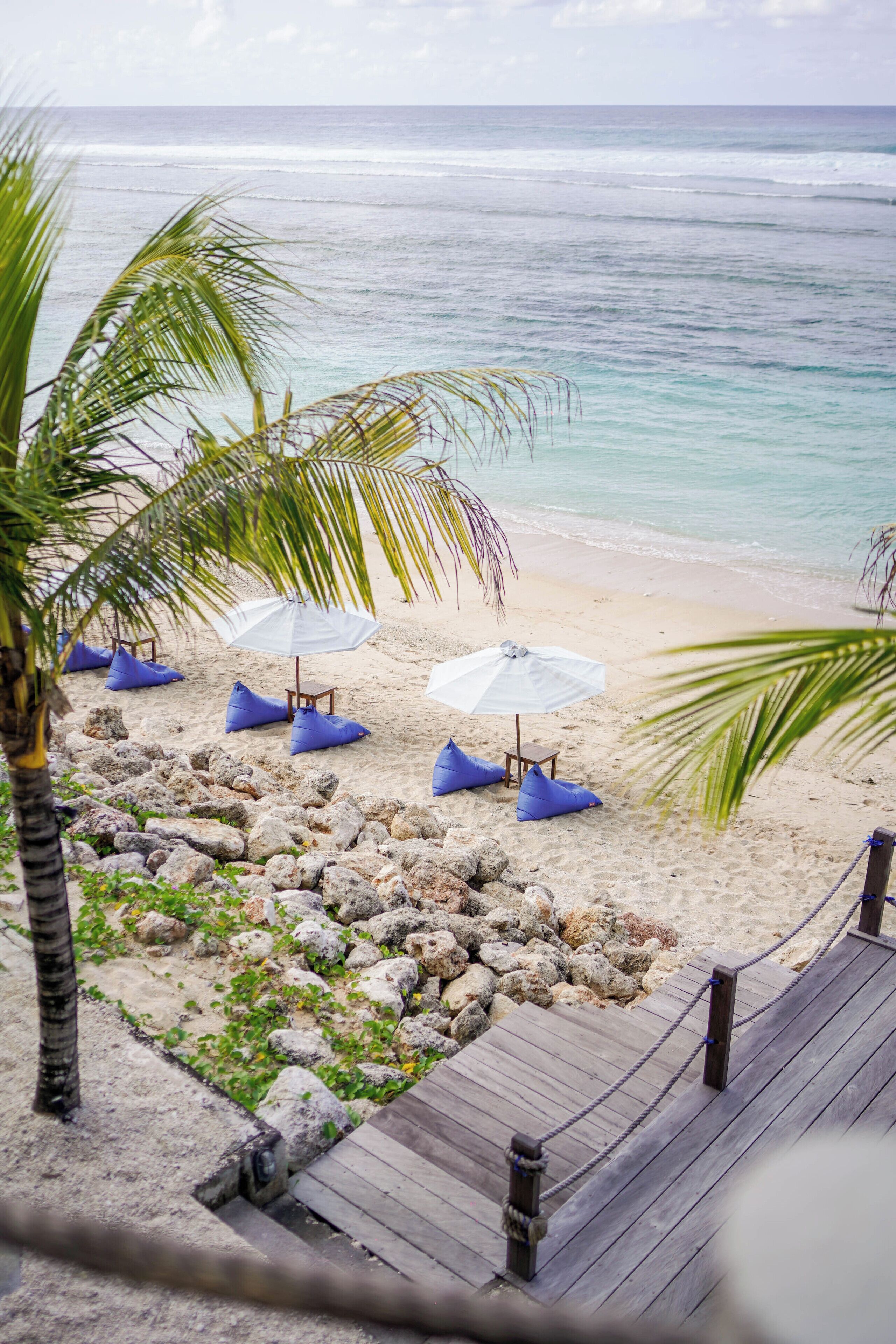 A beach with a wooden deck, white umbrellas, blue cushions, and a palm tree.