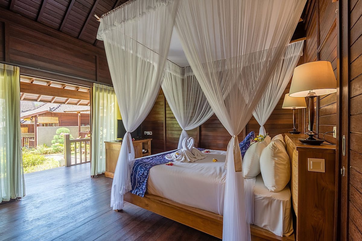 A four-poster canopy bed with a wooden headboard, bedside lamps, and a view of a garden through an open door.