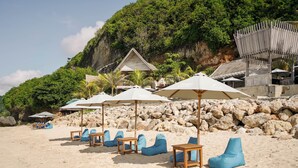 A beach with lounge chairs and umbrellas, a wooden structure, and a rocky cliff with greenery. - Puri Pandawa Resort (Kutuh)