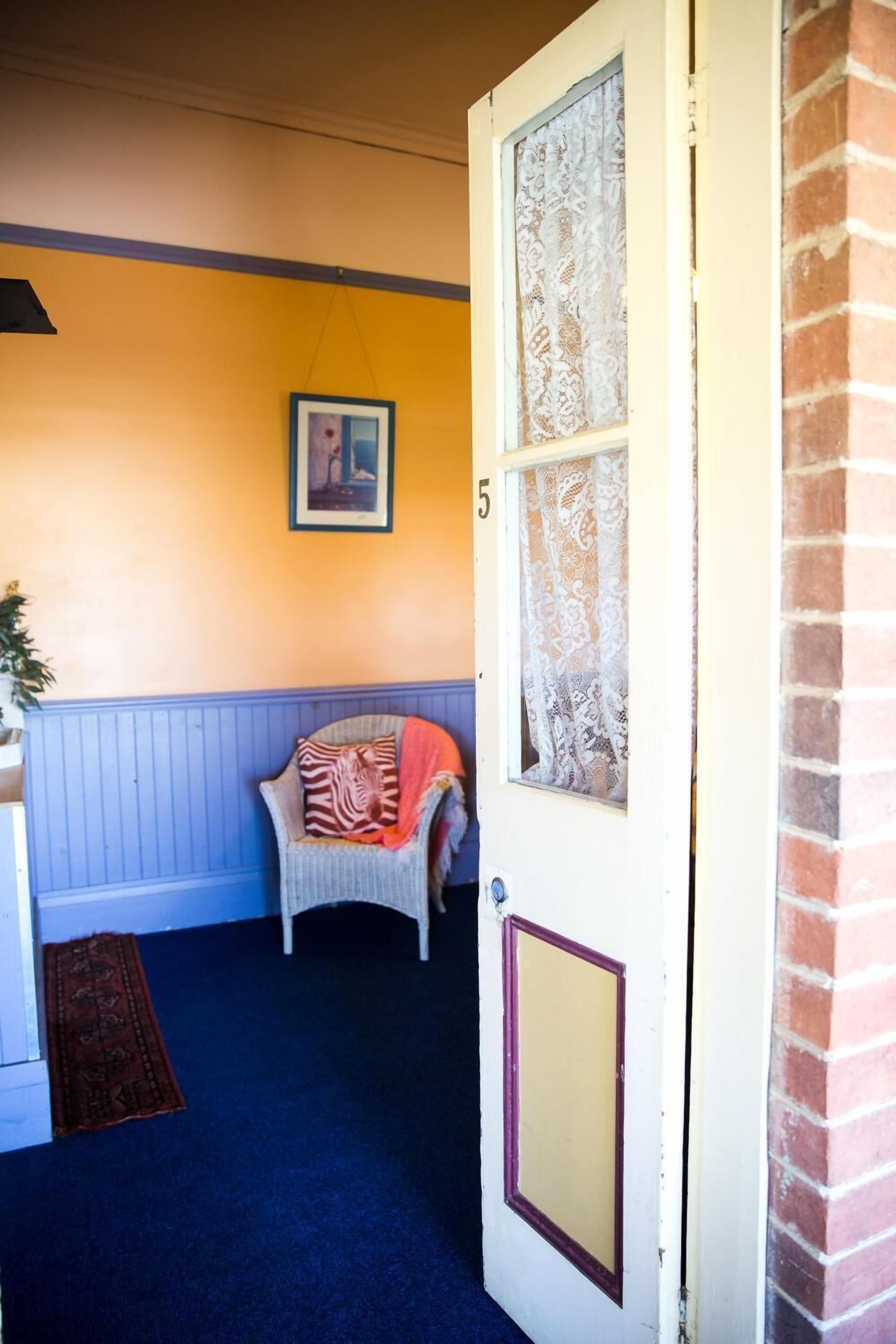 Una habitación con alfombra azul, una puerta blanca con un número, cortina de encaje y un cuadro enmarcado en la pared.